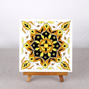 May include: A white ceramic tile with a black, yellow, and orange floral design. The tile is on a wooden easel.