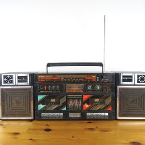 Könnte beinhalten: Ein schwarzer Vintage-Boombox mit silbernen Akzenten, mit einem Radiowählrad, einem Kassettenspieler und Lautsprechern. Die Boombox hat einen Tragegriff und eine Antenne. Sie steht auf einer Holzoberfläche.