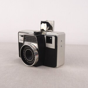 May include: A silver and black Argus Automat 35mm film camera with a built-in flash. The camera has a black lens with a silver ring and the words "Argus Cintar" printed on it.