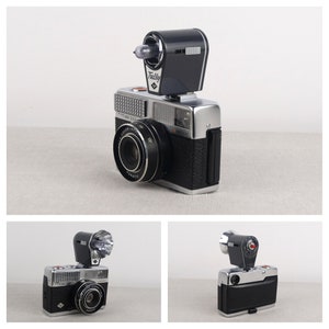 May include: A silver and black 35mm film camera with a flash attachment. The camera has a lens with a black ring around it and the text "OPTIMA 503" on the front. The flash attachment is black and has the text "Tully" on it.