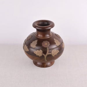 Puede incluir: Un jarrón de madera marrón oscuro con cuerpo redondo y base acampanada. El jarrón está adornado con diseños florales y de hojas de latón. El jarrón tiene una amplia abertura en la parte superior y es adecuado para exhibir flores o como objeto decorativo.