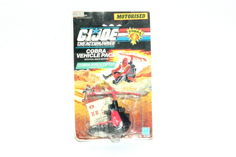 May include: A red and black Cobra Gyrocoptor toy with a pull-back motor. The toy is packaged in a cardboard box with a picture of the toy and the text "GI JOE THE ACTION FORCE COBRA VEHICLE PACK WITH PULL BACK MOTOR COBRA GYROCOPTER MOTORISED COBRA".