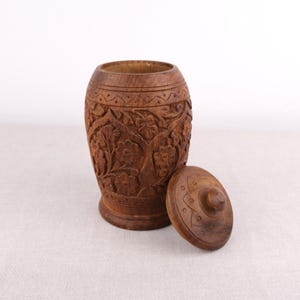 May include: A handcrafted wooden container with a lid. The container is cylindrical with intricate floral carvings. The lid is round with a central knob and matching carved details. The wood has a warm brown tone.