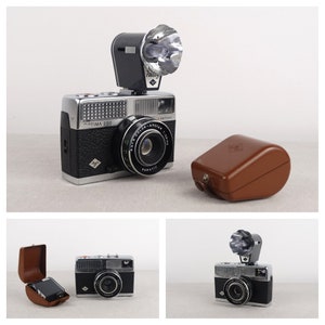 May include: A silver and black 35mm film camera with a built-in flash and a brown leather case. The camera has a lens labeled "Auta Color-Apotar 1:2.8" and "Paratic". The camera is labeled "Optima 200".