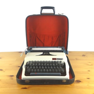 May include: A white vintage Erika typewriter with a black case. The case is open and the typewriter is inside. The typewriter has a black keyboard with white keys and a red label that says "Erika".