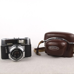 May include: A silver and black Voigtlander Vito B camera with a brown leather case. The camera has a silver lens with the text "Voigtlander Color Skopar 2/50" printed on it.