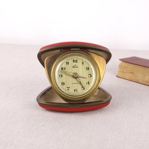 May include: A gold-toned alarm clock with a white face and black hands. The clock is in a red leather case and is open.