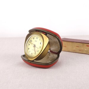May include: A gold-toned alarm clock with a white face and black hands. The clock is in a red leather case and is open.