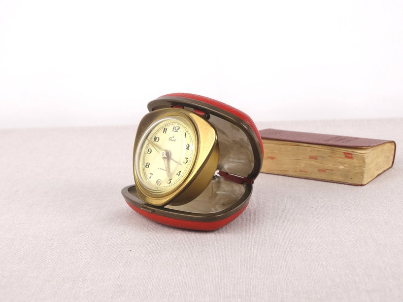 May include: A gold-toned alarm clock with a white face and black hands. The clock is in a red leather case and is open.