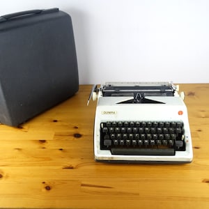 May include: A white vintage Olympia typewriter with a black keyboard and a black carrying case. The typewriter has a red button on the right side.