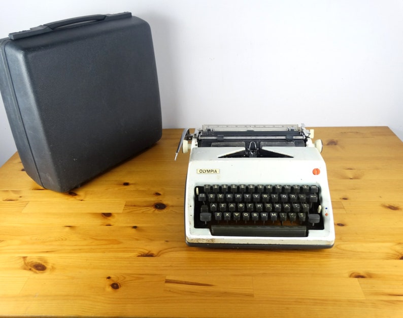 Puede incluir: Una m&aacute;quina de escribir Olympia vintage blanca con un teclado negro y una maleta de transporte negra. La m&aacute;quina de escribir tiene un bot&oacute;n rojo en el lado derecho.