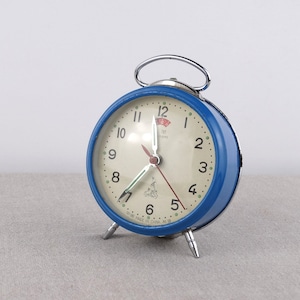 Könnte beinhalten: Ein blauer Wecker mit weißem Zifferblatt und schwarzen Zahlen. Die Uhr hat einen silbernen Griff und silberne Füße. Das Zifferblatt der Uhr hat einen roten Sekundenzeiger und einen grünen Minutenzeiger. Das Zifferblatt der Uhr hat auch den Text "MADE IN CHINA" unten.