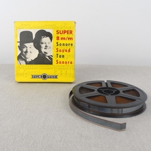 May include: A vintage film reel and box. The yellow box features a black and white image of two men and the text "SUPER 8 m/m Sonore Sound Ton Sonoro". The film reel is black and brown.