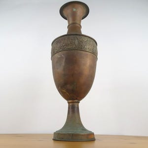 May include: A large, antique copper vase with an ornate design. The vase has a wide base and a narrow neck, and it is decorated with a band of embossed flowers and leaves.