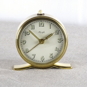May include: A vintage gold-toned alarm clock with a white face and black Roman numeral hour markers. The clock has a gold-toned base and the brand name "Albau" is printed on the face.
