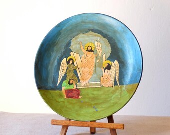 Pode incluir: Um prato redondo de cerâmica com uma cena pintada de uma figura religiosa com asas, cercada por outras duas figuras com asas. A cena é pintada em um estilo colorido com tons azuis, verdes, amarelos e vermelhos.
