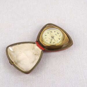 May include: A gold-colored, triangular-shaped alarm clock with a white face and black hands. The clock is open and the inside of the case is red.