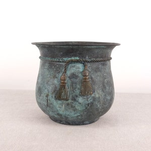 Può includere: Un vaso di metallo verde con un design a corda e nappe intorno alla parte superiore.