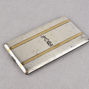 Könnte beinhalten: Ein rechteckiges Silber- und Gold-Etui mit einem Monogramm "M" in der Mitte. Das Etui hat zwei goldene Streifen, die horizontal über die Oberfläche verlaufen.