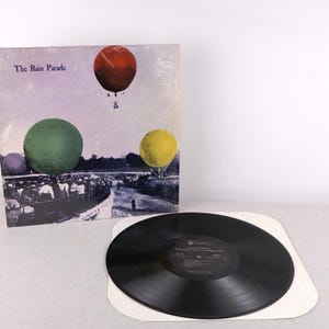Puede incluir: Un álbum de vinilo titulado "The Rain Parade" con una portada colorida con globos aerostáticos. El disco de vinilo negro está sobre una funda de papel blanca. La portada del álbum tiene un fondo morado y blanco.