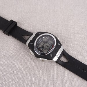 May include: Black digital wristwatch with a silver face and black rubber band. The watch face displays the time, date, and day of the week. The watch brand is "WJAGA".