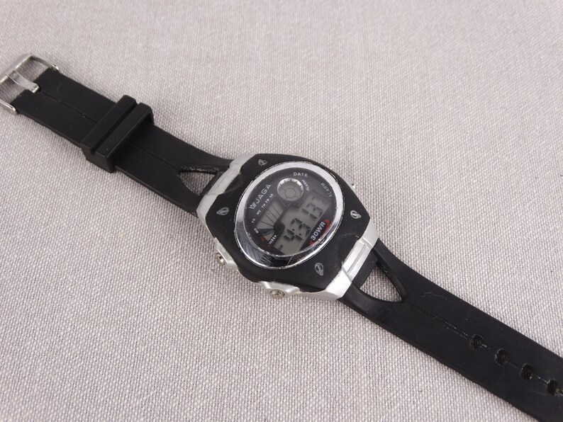 May include: Black digital wristwatch with a silver face and black rubber band. The watch face displays the time, date, and day of the week. The watch brand is "WJAGA".
