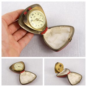 May include: A gold-toned, triangular alarm clock with a white face and black hands. The clock is open and the inside of the case is lined with white fabric. The clock is sitting on a white surface.