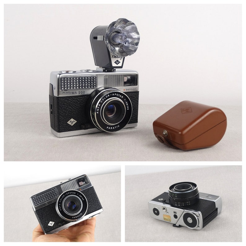 May include: A black and silver 35mm film camera with a flash attachment. The camera has a lens labeled "Agfa Color-Apotar 1:2.8/40" and the text "Optima 200" on the front. The camera is sitting on a white surface.