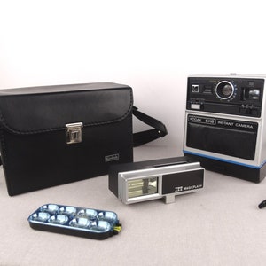 Puede incluir: Una funda de cámara de cuero negro con un cierre plateado y la palabra "Kodak" en la parte delantera. Una cámara instantánea Kodak Ek6 plateada y negra con una franja azul y el texto "Kodak Ek6 Instant Camera". Una unidad de flash plateada y negra con el texto "ITT Magicflash". Una caja de plástico negra con ocho bombillas de flash azules.