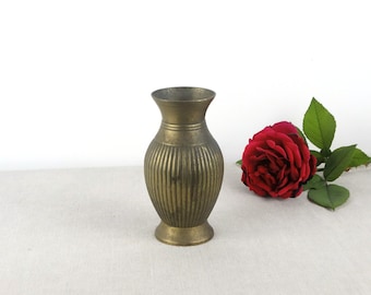 May include: A small, gold-colored metal vase with a ribbed design. The vase is sitting on a white surface with a red rose next to it.