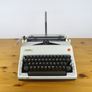 Puede incluir: Una m&aacute;quina de escribir Olympia vintage blanca con un teclado negro y un bot&oacute;n rojo. La m&aacute;quina de escribir est&aacute; sobre una superficie de madera.