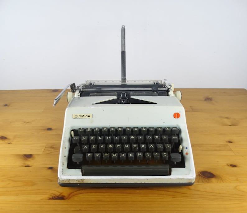 Puede incluir: Una m&aacute;quina de escribir Olympia vintage blanca con un teclado negro y un bot&oacute;n rojo. La m&aacute;quina de escribir est&aacute; sobre una superficie de madera.