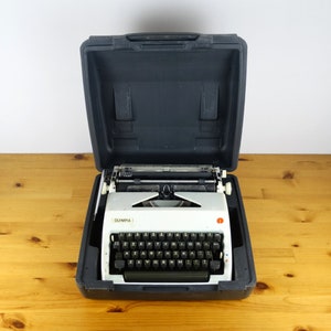 Puede incluir: Una m&aacute;quina de escribir Olympia blanca con un teclado negro y una maleta de transporte negra. La m&aacute;quina de escribir est&aacute; abierta y lista para ser usada.