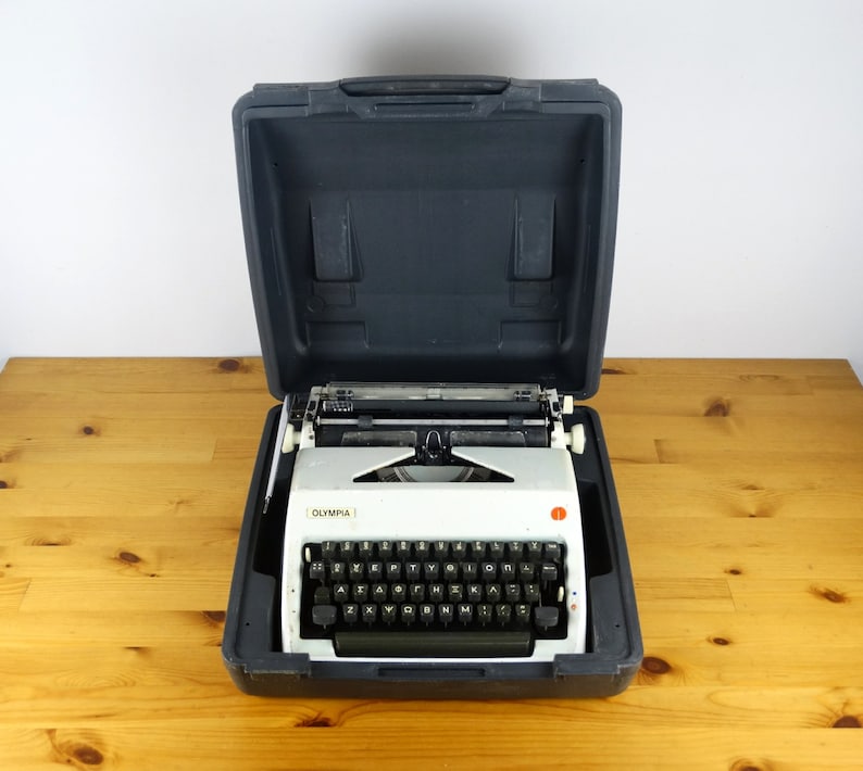 Puede incluir: Una m&aacute;quina de escribir Olympia blanca con un teclado negro y una maleta de transporte negra. La m&aacute;quina de escribir est&aacute; abierta y lista para ser usada.