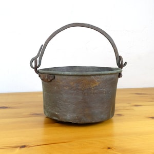 May include: An antique copper pot with a wrought iron handle. The pot has a worn, aged patina and is sitting on a wooden surface.