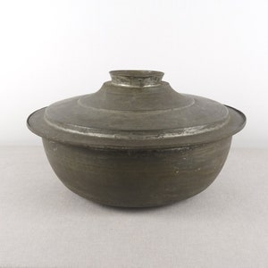 Puede incluir: Una olla redonda de metal vintage con tapa. La olla es de color gris mate y tiene un aspecto ligeramente desgastado.