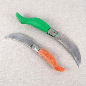 May include: Two small, curved knives with different coloured handles. The knife on the left has an orange handle and the knife on the right has a green handle.