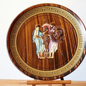 May include: A round wooden plate with a gold trim and a raised image of three figures in a Greek style. The figures are painted in shades of blue, white, and pink.
