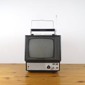 May include: A black and silver portable television with a built-in antenna. The television has a small screen and a dial for channel selection. The television is on a wooden surface.
