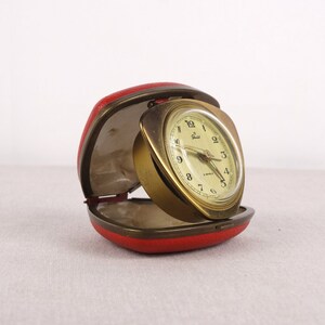 May include: A gold-colored alarm clock with a white face and black hands. The clock is in a red leather case.