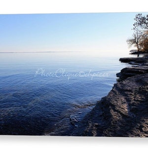 Op de afbeelding: Een foto van een rotsachtige kustlijn met een kalm blauw meer op de achtergrond. De lucht is lichtblauw en het water is diepblauw. De rotsen zijn donkergrijs en het water reflecteert de lucht.