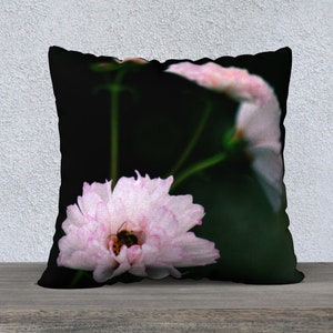 May include: A decorative pillow featuring a close-up of a light pink flower with a bee inside. The flower has a ruffled edge and is set against a dark green and black background. The pillow is square and rests on a wooden surface.