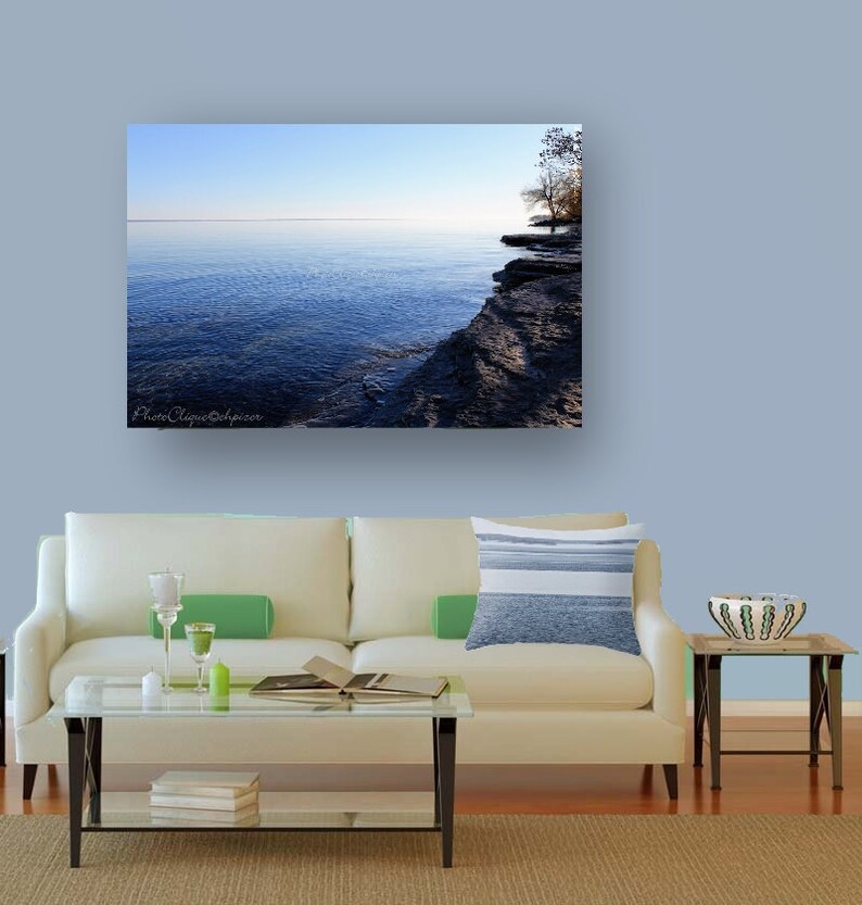 Op de afbeelding: Een foto van een kalm blauw meer met een rotsachtige kustlijn en een enkele boom aan de andere kant. De lucht is lichtblauw en het water reflecteert de lucht.