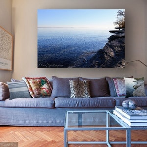 Op de afbeelding: Een woonkamerinterieur met een grijze bank en een glazen salontafel. De bank heeft verschillende kussens met patronen. Een grote ingelijste foto van een meer en een rotsachtige kustlijn hangt aan de muur boven de bank.