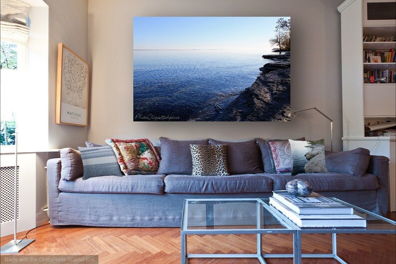 Op de afbeelding: Een woonkamerinterieur met een grijze bank en een glazen salontafel. De bank heeft verschillende kussens met patronen. Een grote ingelijste foto van een meer en een rotsachtige kustlijn hangt aan de muur boven de bank.