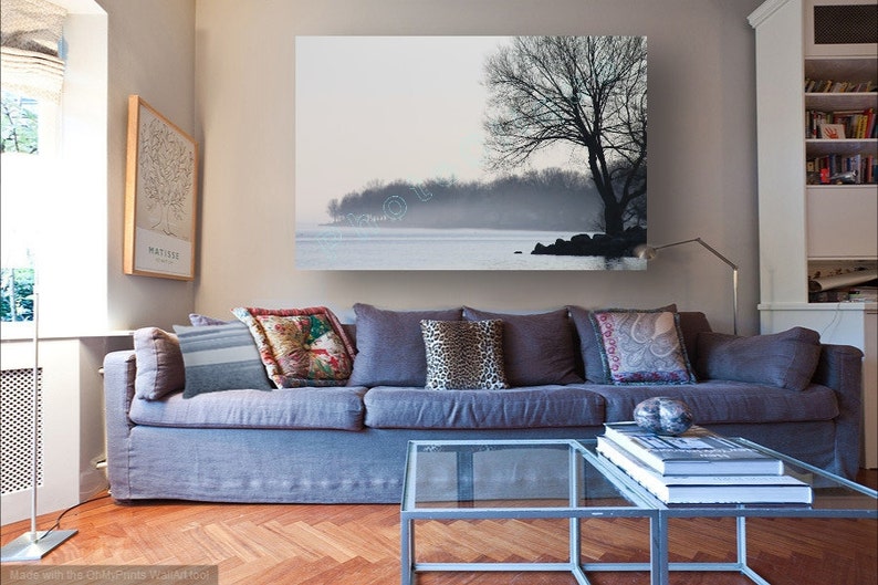 May include: A gray sofa with pillows in a living room setting. A large framed print of a tree and a lake hangs on the wall above the sofa. A glass coffee table sits in front of the sofa with a stack of books on top.