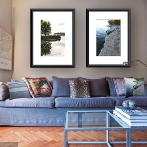 May include: Two framed landscape photographs of a lake with trees and a rocky shoreline hang on a wall above a gray couch with pillows. A glass coffee table sits in front of the couch with a stack of books and a decorative object.