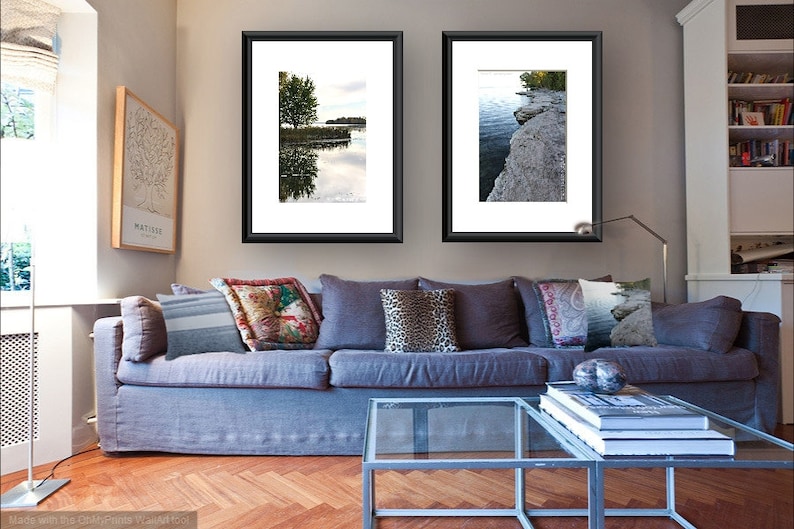 May include: Two framed landscape photographs of a lake with trees and a rocky shoreline hang on a wall above a gray couch with pillows. A glass coffee table sits in front of the couch with a stack of books and a decorative object.