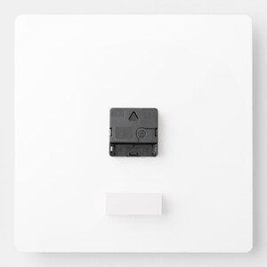 Puede incluir: La parte posterior de un reloj blanco con un mecanismo de reloj negro y un peque&ntilde;o rect&aacute;ngulo blanco.