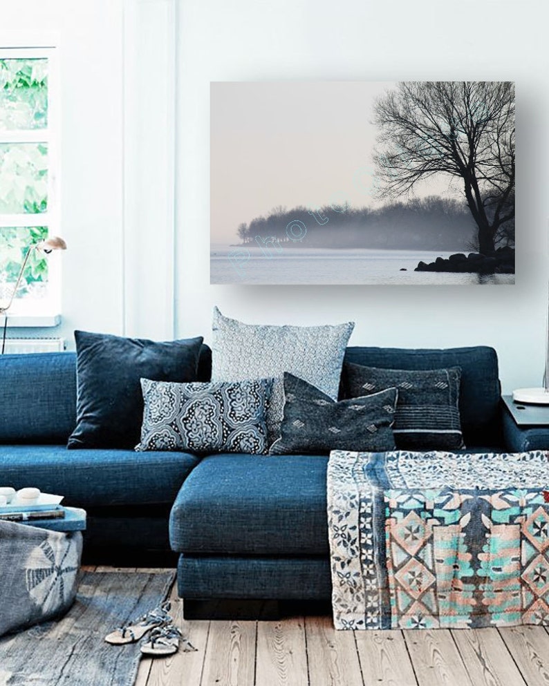 May include: A blue sectional sofa with a variety of patterned throw pillows and a throw blanket. The sofa is in front of a wall with a framed print of a foggy lake and a tree.